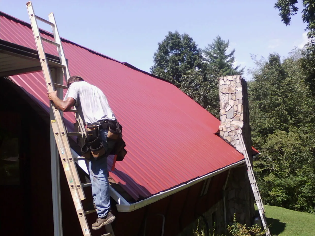 Roofer on a ladder beside a red metal roof and stone chimney — Soffit & Fascia Repair by Roofing Brothers in Evergreen Park